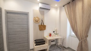 A modern bathroom with a wooden door, a desk with a basket, a clear chair, and a window with beige curtains. - LA DOLCE VITA ROOMS (Procida)