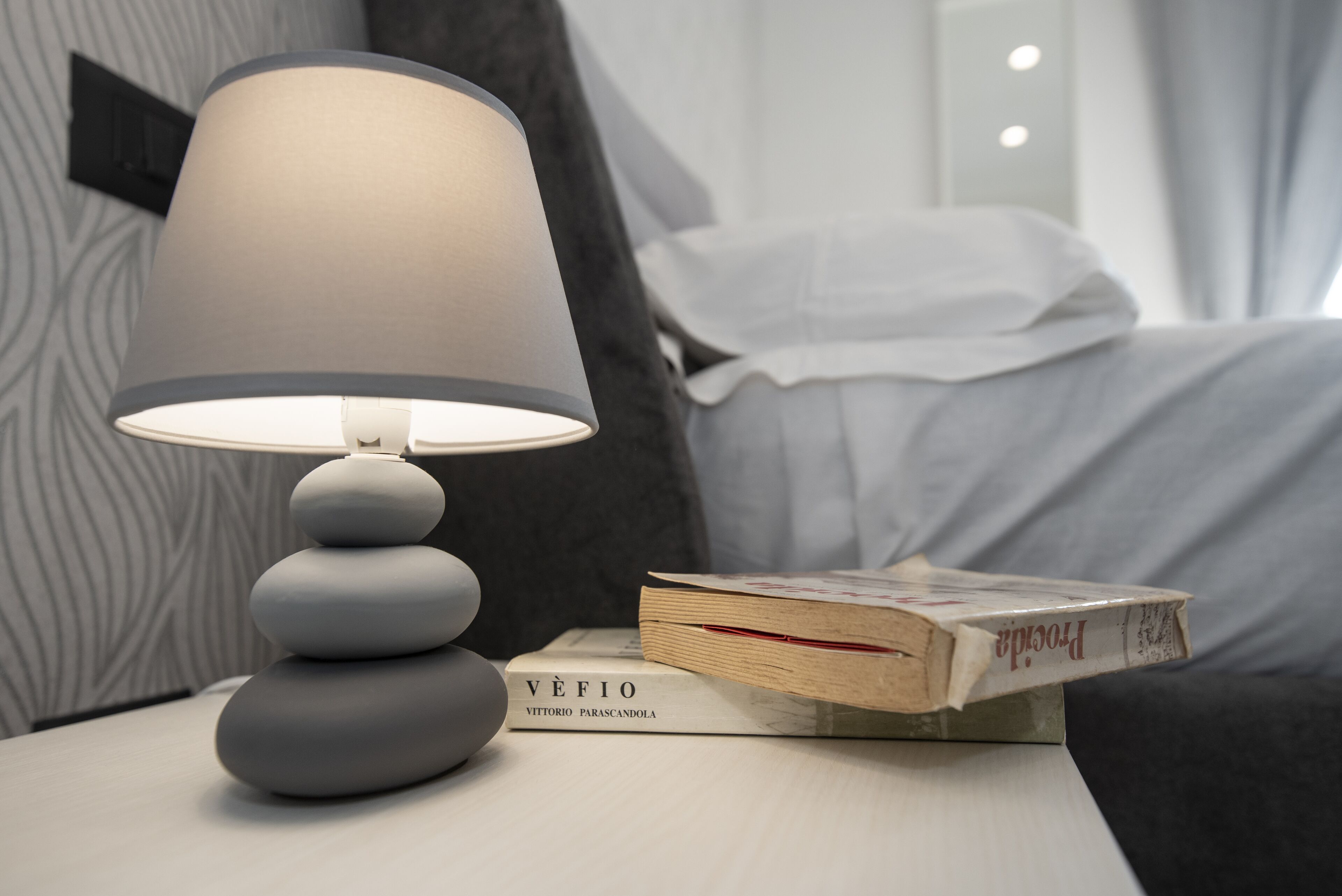 Una mesita de noche con una lámpara, un libro titulado "Véfio" de Vittorio Parascandola, y una cama con ropa blanca de cama.