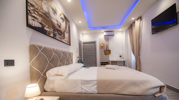 A modern bedroom with a large bed, a bedside table with a lamp, a TV mounted on the wall, and a painting above the bed. - LA DOLCE VITA ROOMS (Procida)