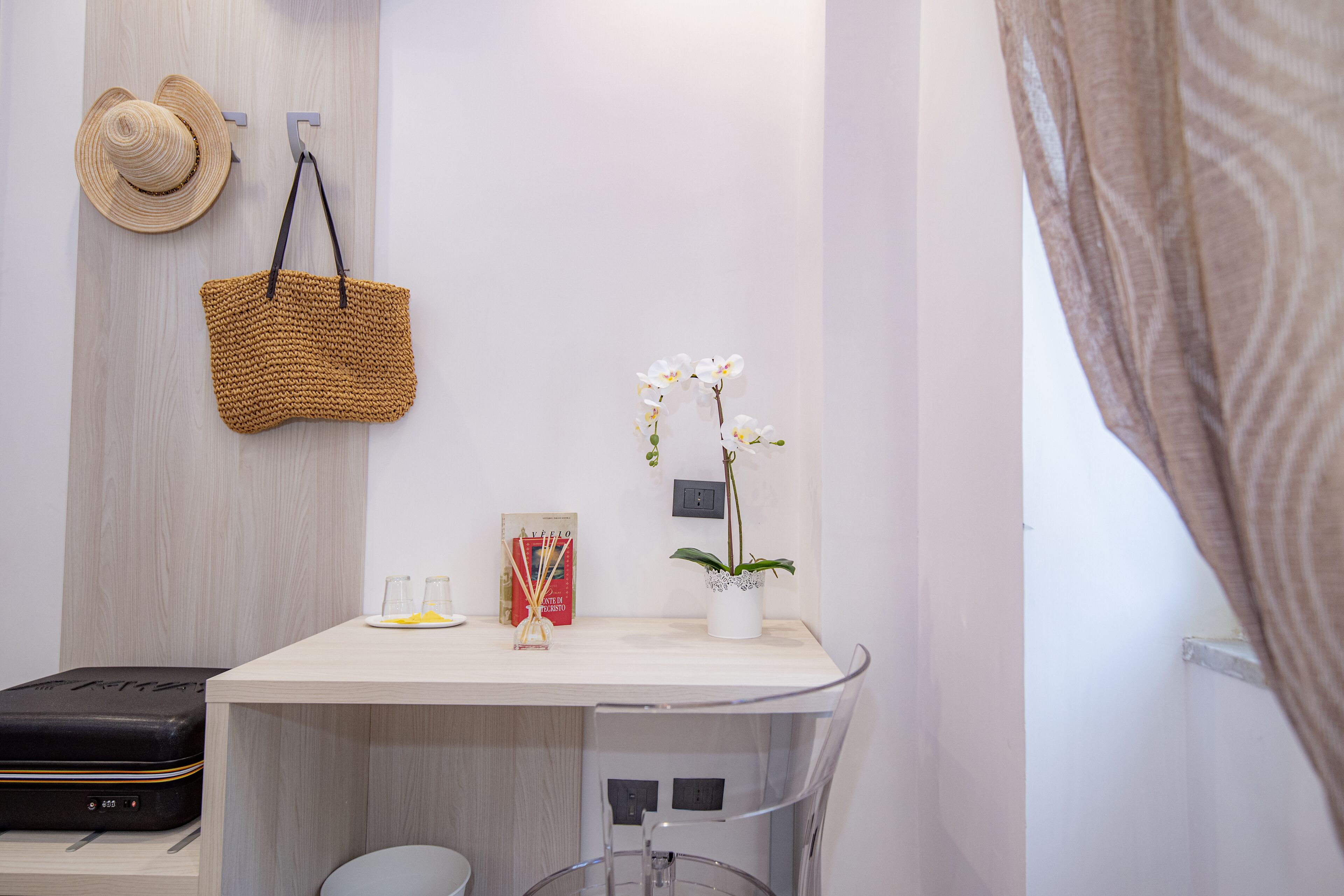 A room with a desk, a suitcase, a hat, a basket, a vase with flowers, and a framed picture.