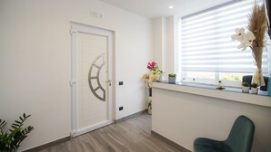 A reception area with a modern design, featuring a white door with a geometric handle, a reception desk with a vase of flowers, and a window with blinds. - LA DOLCE VITA ROOMS (Procida)