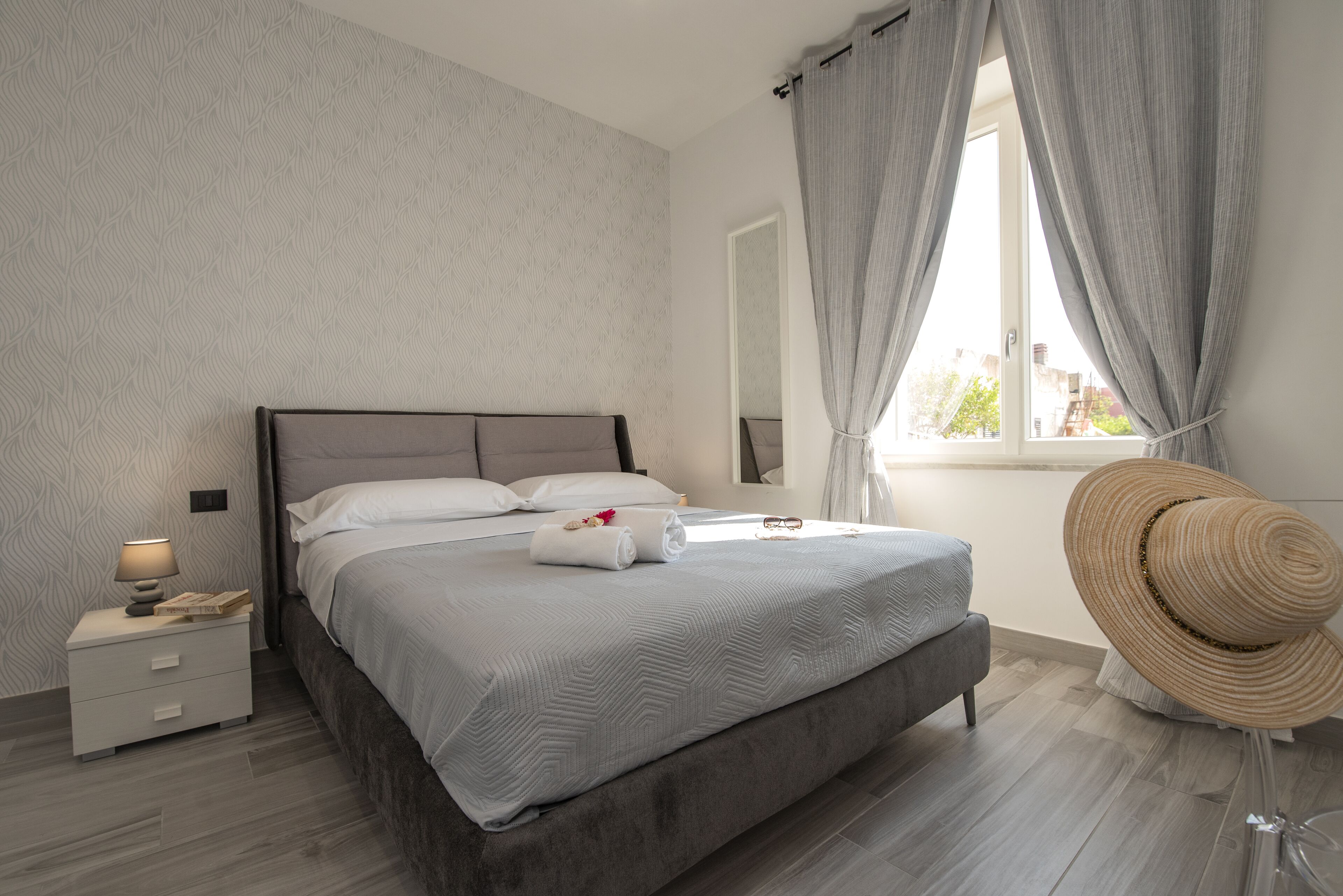 A neatly made bed with a grey headboard, a bedside table with a lamp, and a large window with curtains.