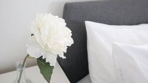 En vit blomma i en genomskinlig glasvas på en vit yta, med en grå sänggavel och en vit kudde i bakgrunden.