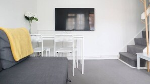 Ett modernt vardagsrum med en grå soffa, en gul pläd, ett vitt skrivbord med två stolar och en platt-TV som är monterad på väggen.