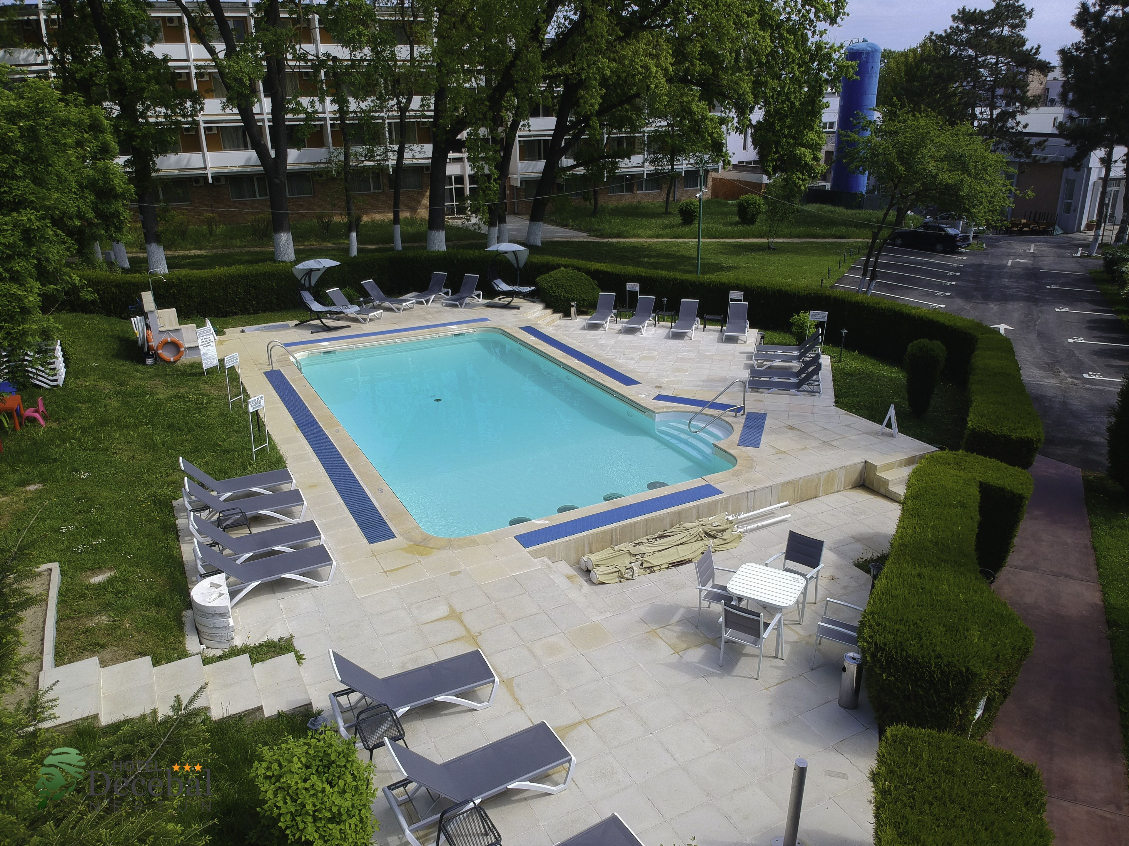 A rectangular pool with a blue border, surrounded by lounge chairs and umbrellas, set in a landscaped area with trimmed hedges and trees.