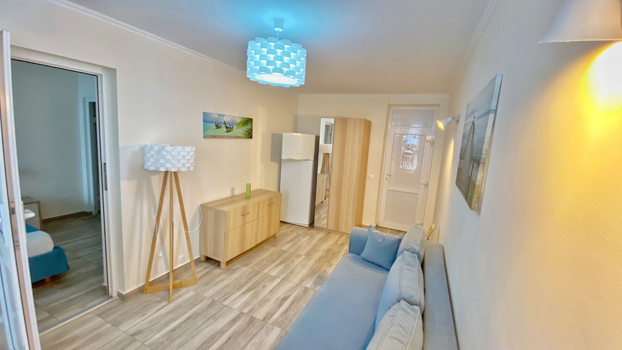 A compact living space with a wooden floor, a light-colored sofa, a small cabinet, a white refrigerator, and a ceiling light fixture.