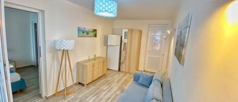 A compact living space with a wooden floor, a light-colored sofa, a small cabinet, a white refrigerator, and a ceiling light fixture.