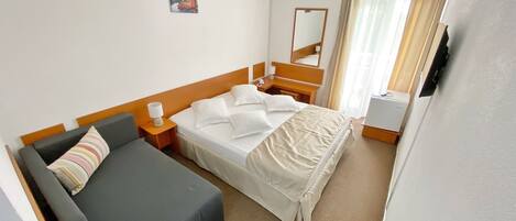 Habitación de hotel con cama, sofá, mesita de noche con lámpara, espejo y ventana con cortinas.