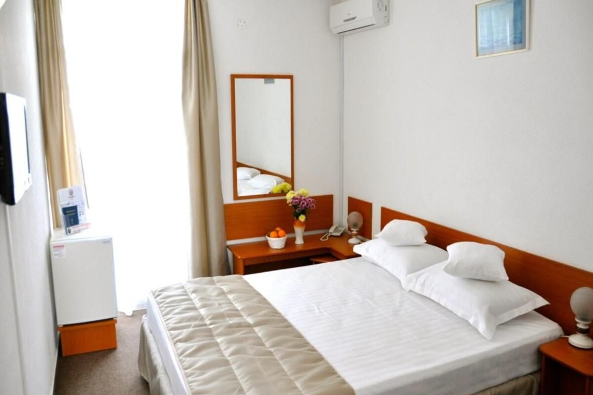 A hotel room with a bed, a bedside table with a lamp, a mirror, a small desk with a phone, and an air conditioning unit.