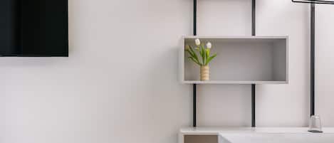 A modern, minimalist room with white walls, a flat-screen TV mounted on the wall, and a series of white shelves with a vase of white flowers on one of them.