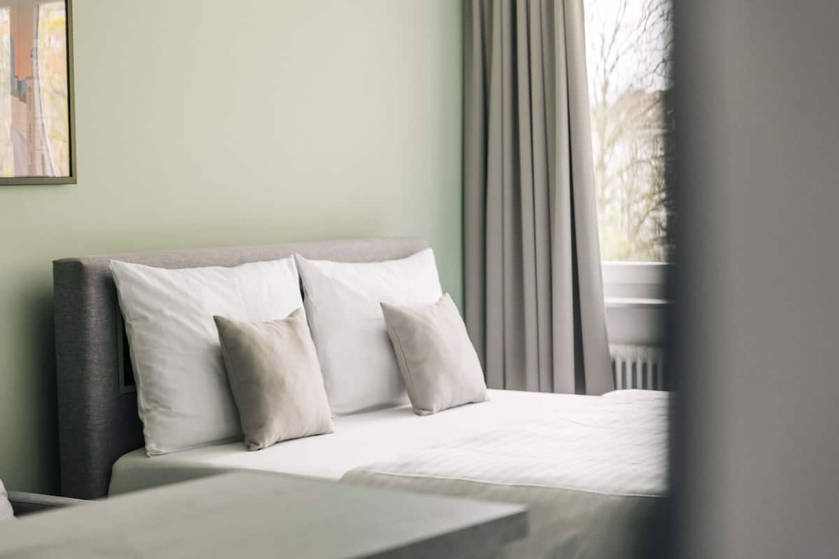 Ein ordentlich bezogenes Bett mit weißen Leinen und mehreren Kissen, ein grauer Kopfteil und ein Blick auf ein Gebäude durch ein Fenster mit Vorhängen.