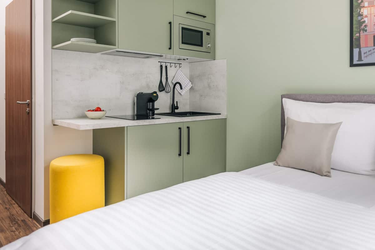 Ein kleines Zimmer mit einem Bett, einer kompakten Küchenzeile inklusive Mikrowelle, Spüle und einem gelben Hocker.