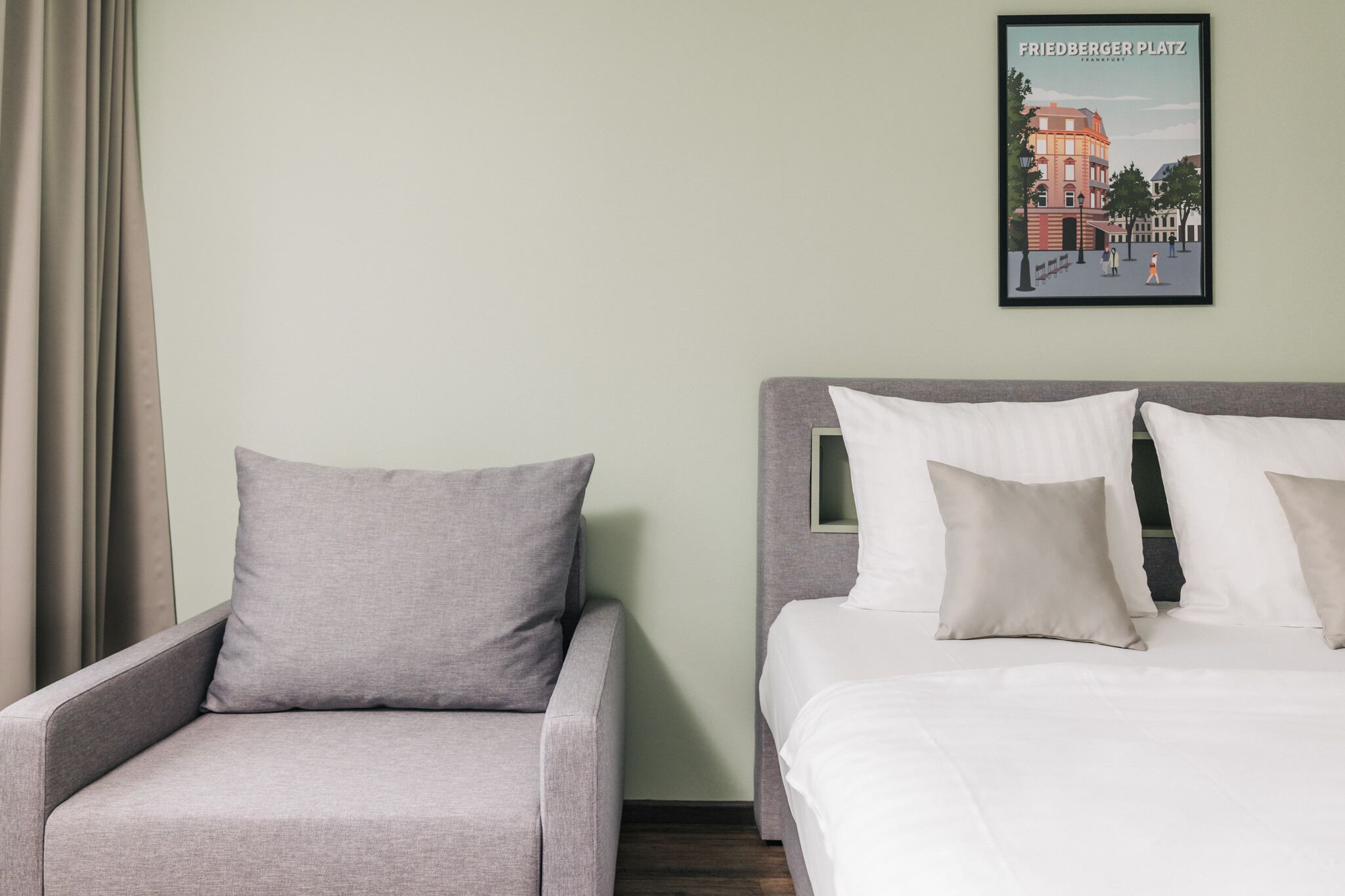 Un cuarto de hotel con un sofá gris, una cama con ropa blanca y un cartel con un paisaje urbano enmarcado en la pared.