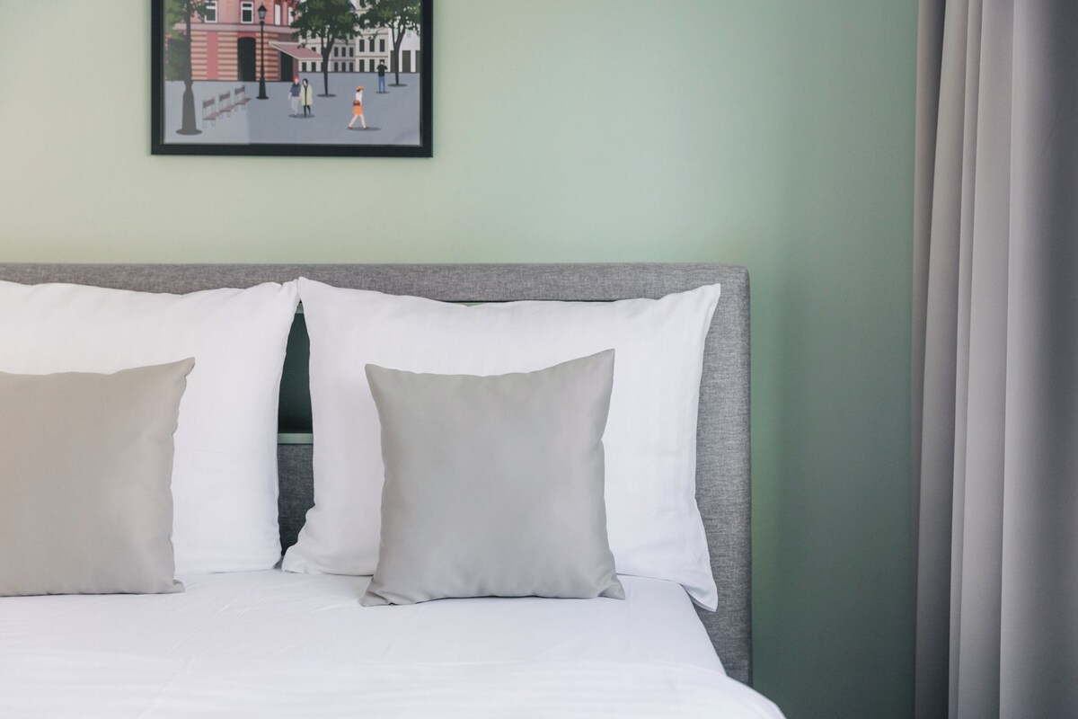 A bed with white and beige pillows, a grey headboard, and a framed picture of a street scene on the wall.