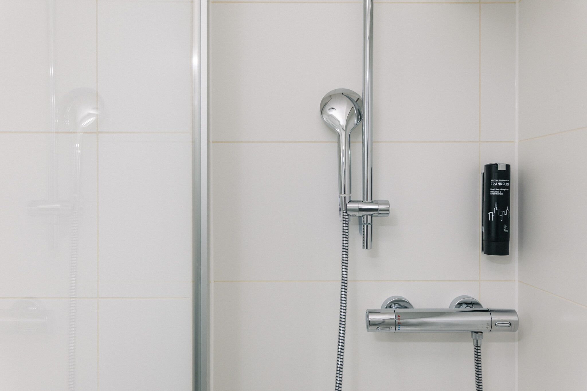 A modern bathroom with a glass shower enclosure, a handheld showerhead, a wall-mounted faucet, and a black control unit.