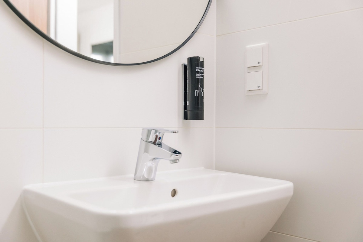 Ein weißes Waschbecken mit chromfarbenem Wasserhahn, einem schwarzen Seifenspender und einem runden Spiegel an der Wand.
