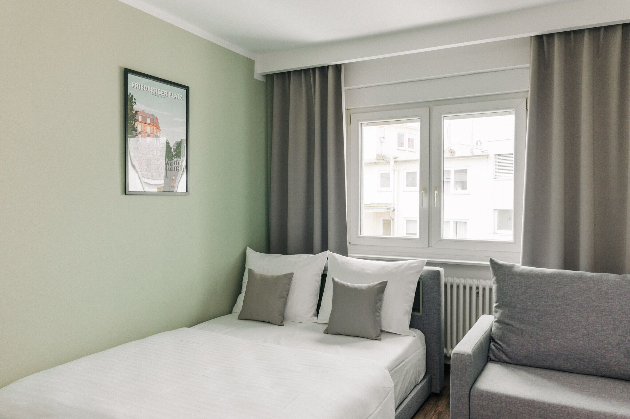 Ein Hotelzimmer mit einem Bett, einem grauen Sofa, einem Fenster mit Vorhängen und einem gerahmten Bild an der Wand.