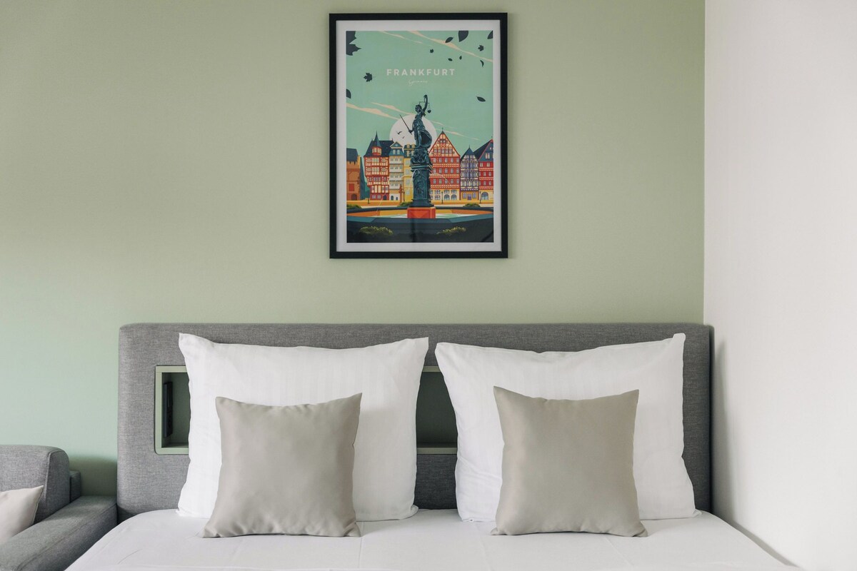 Ein Hotelzimmer mit einem Bett, zwei Kissen, einem gerahmten Stadtbild-Poster und einem grauen Kopfteil.