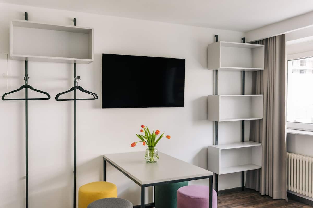 Ein modernes Hotelzimmer mit Flachbildfernseher, einem kleinen Tisch mit Blumenvase und einer Sitzecke mit farbenfrohen Poufs.