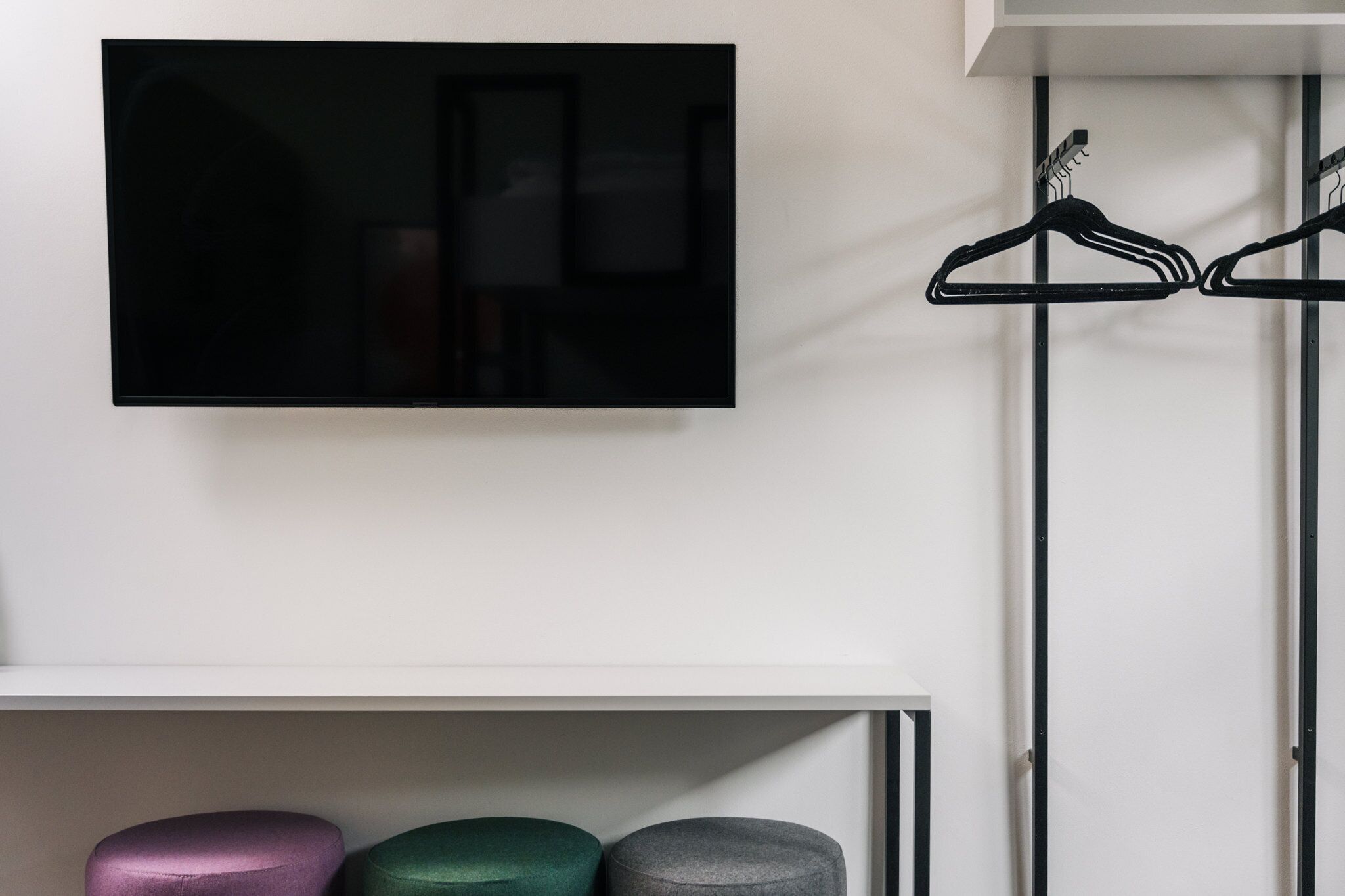 A television mounted on a wall, a clothing rack with hangers, and three ottomans in different colors.