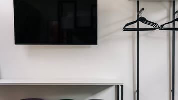 A television mounted on a wall, a clothing rack with hangers, and three ottomans in different colors.