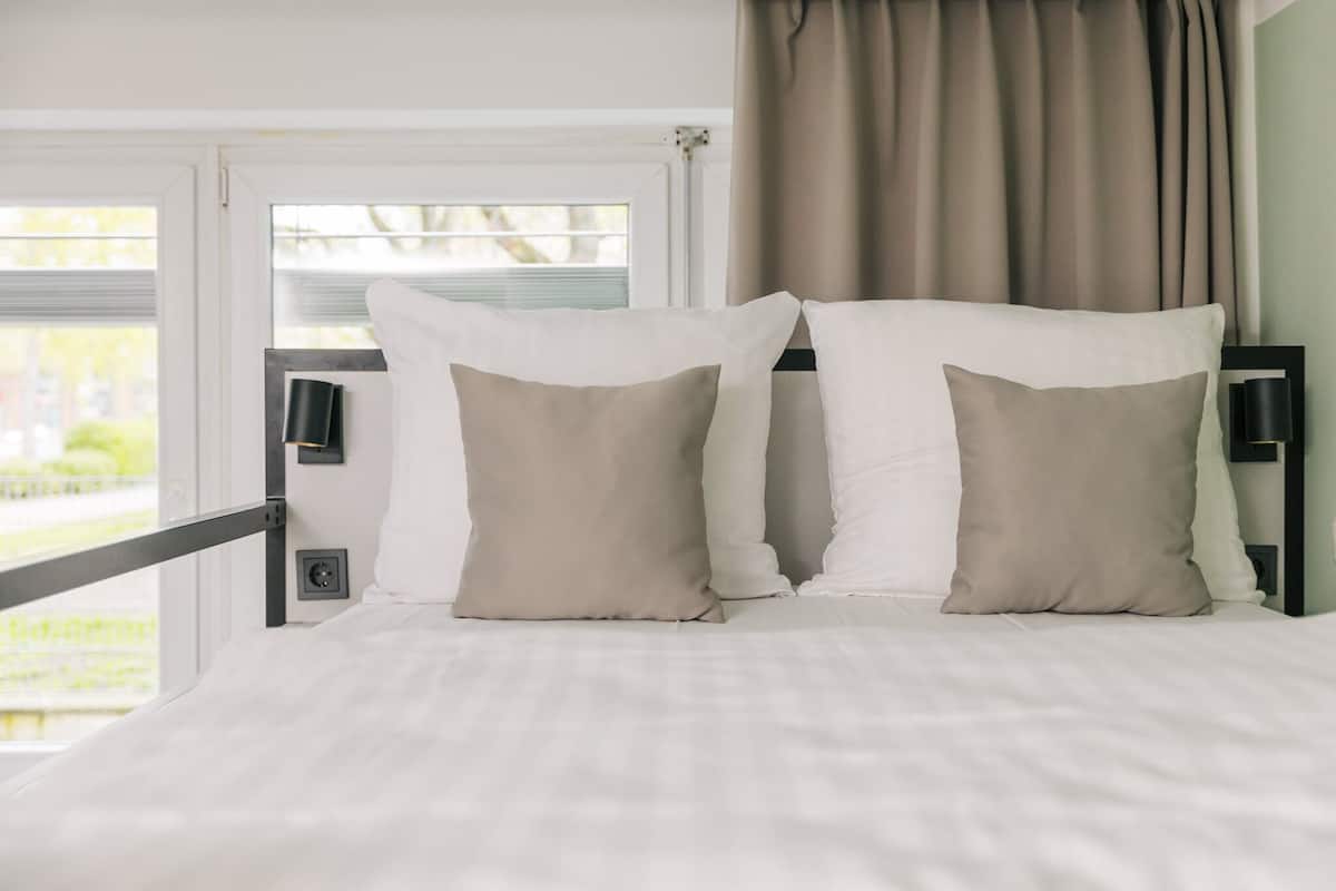 Ein Bett mit weißer Bettwäsche, zwei beigen Kissen, einem schwarzen Kopfteil und einem Fenster mit Vorhängen.