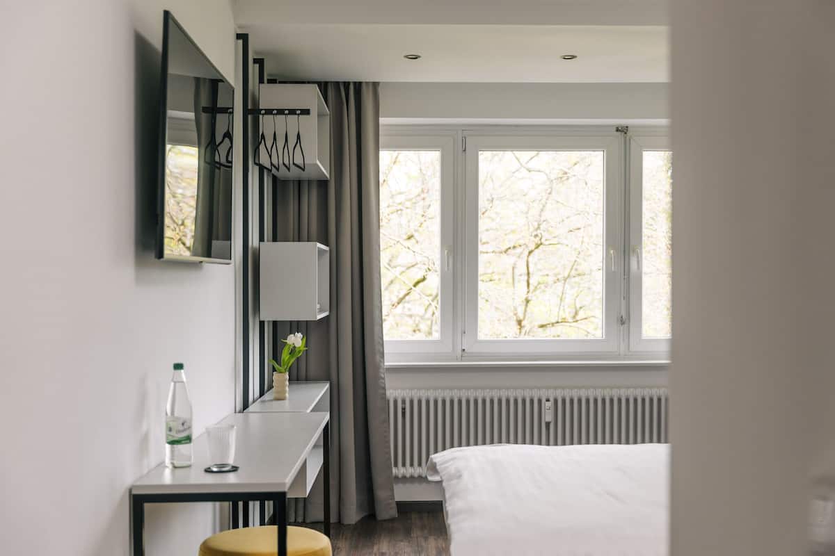 Ein modernes Hotelzimmer mit Bett, Schreibtisch mit Stuhl, Fenster mit Vorhängen und Heizkörper.