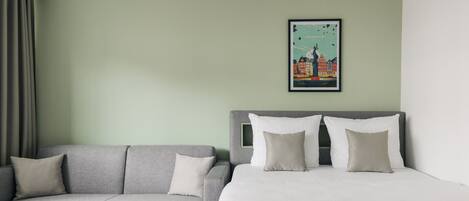 Une chambre d’hôtel avec un canapé gris, un lit avec une literie blanche et une affiche encadrée représentant un paysage urbain accrochée au mur.