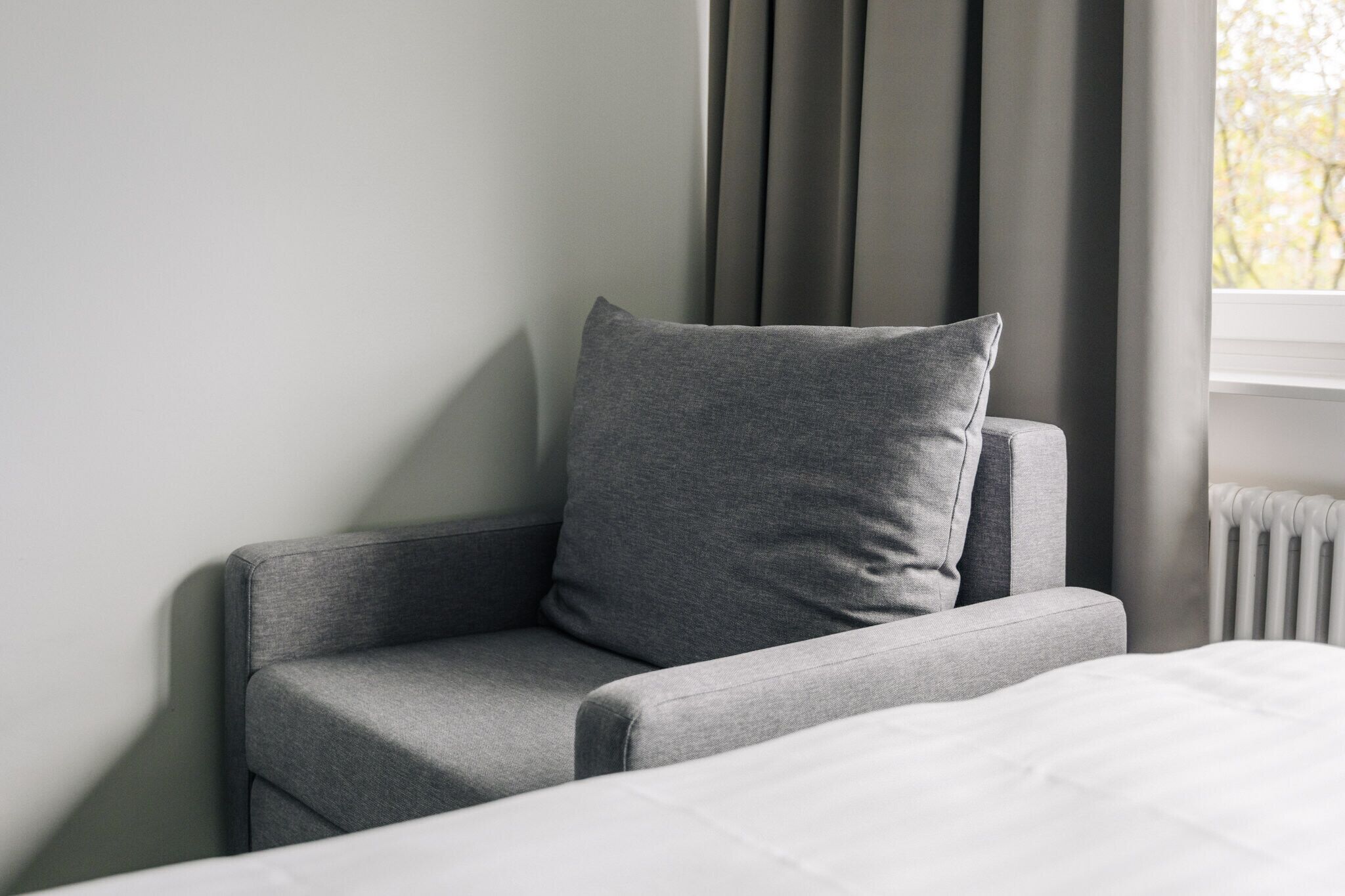 A modern room with a grey armchair, a white bed, and a window with curtains.