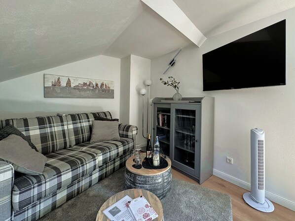 Ein Wohnzimmer mit einem karierten Sofa, einem runden Couchtisch mit Papieren, einer Stehlampe, einem Bücherregal und einem Heizlüfter.