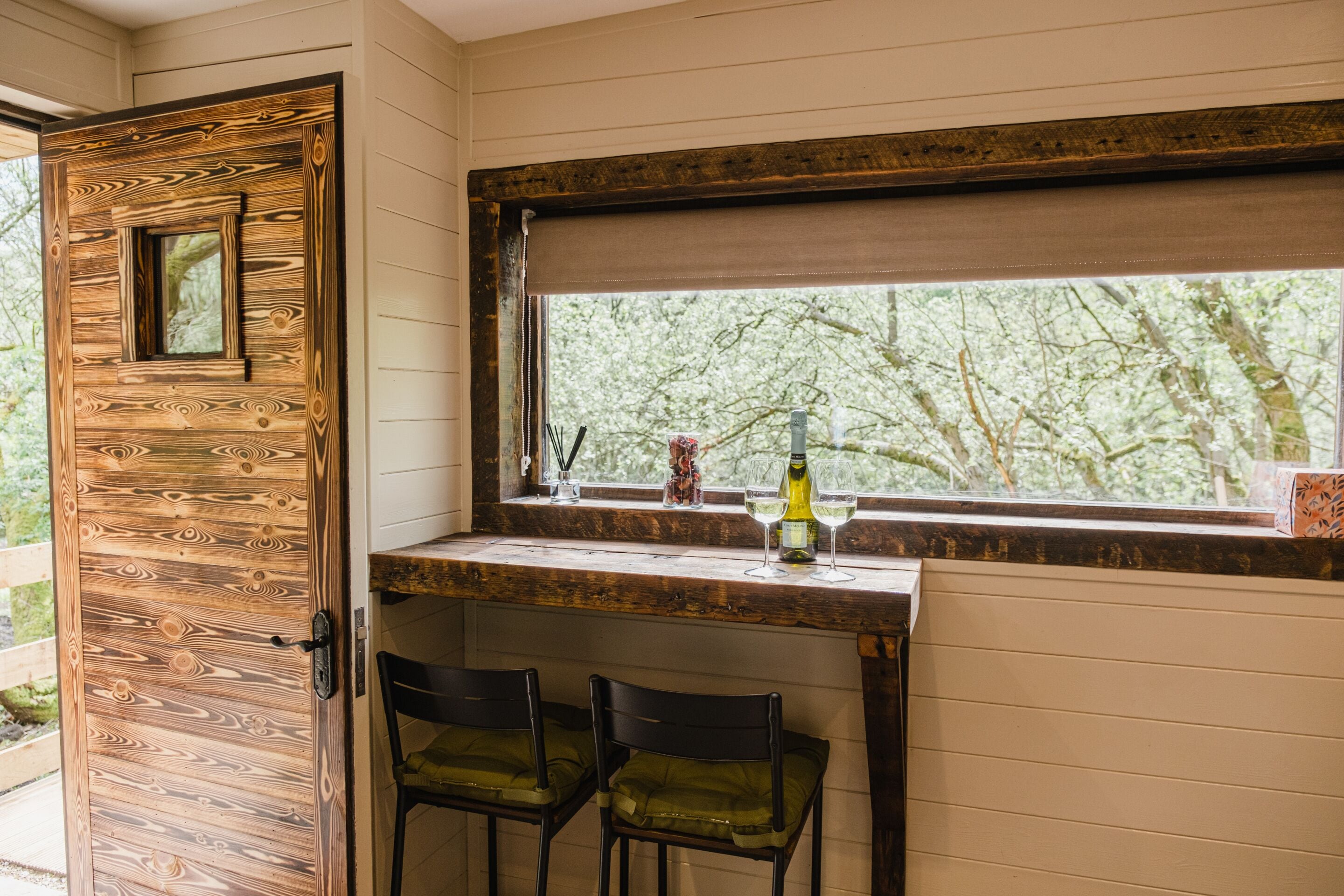 Una habitación con puerta de madera, ventana con vista a árboles, zona de bar con dos sillas y una botella en la encimera.