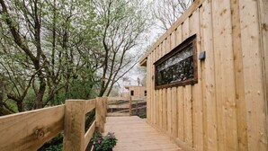 Un sentier en bois mène à un bâtiment doté d’une fenêtre, le tout étant entouré d’arbres.