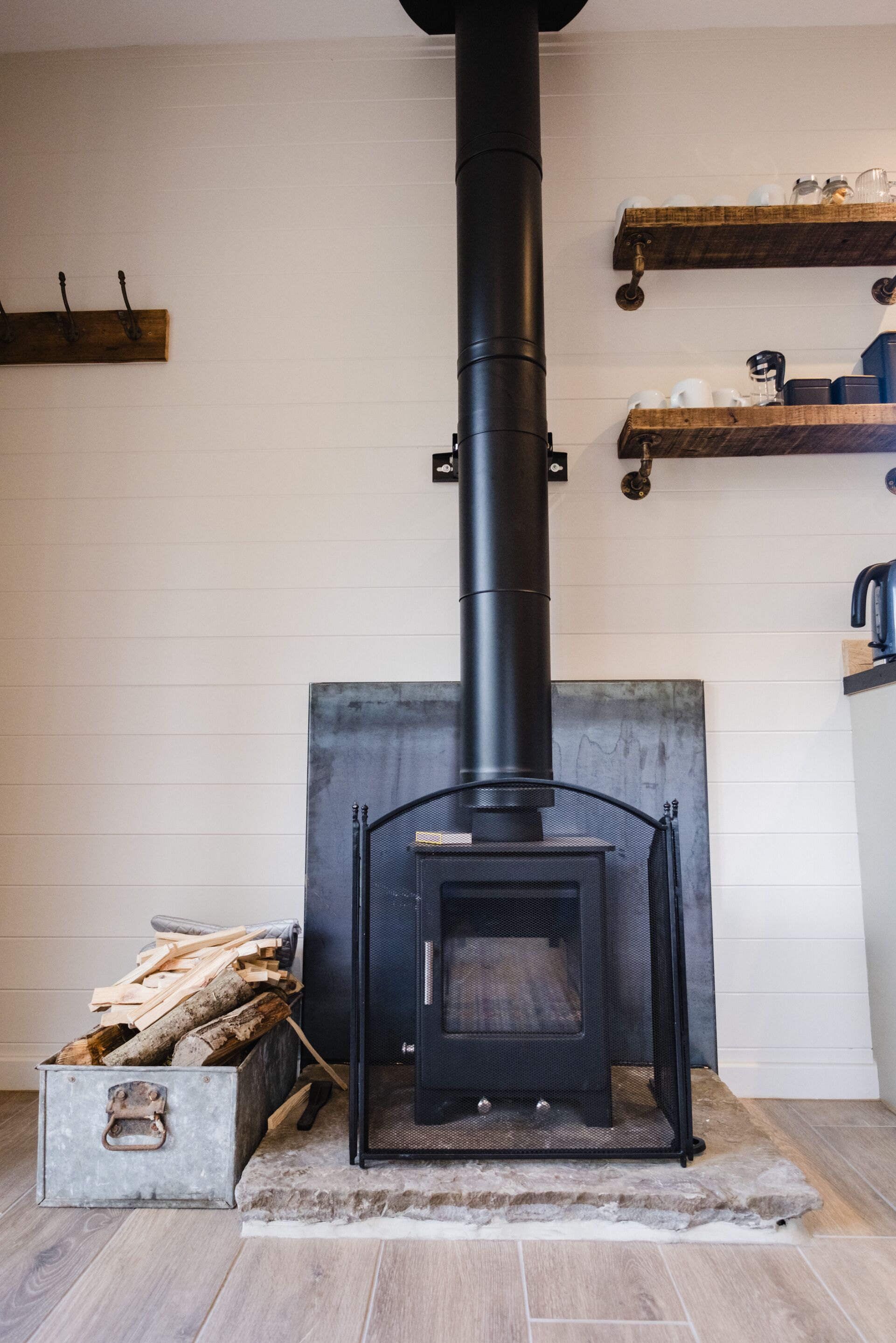 Una estufa de leña con chimenea, un depósito para leña y un estante con utensilios de cocina.
