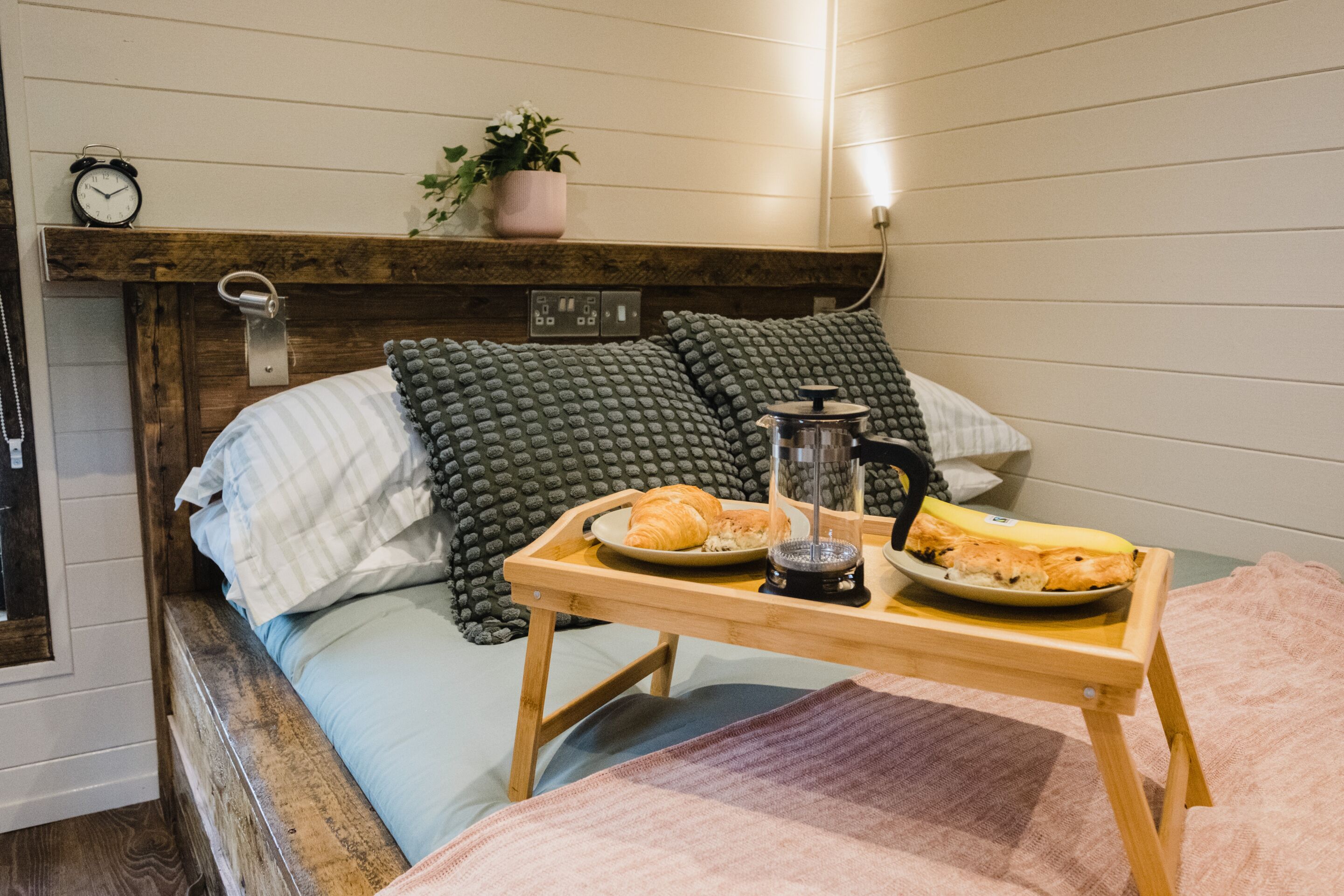 Un dormitorio acogedor con cabecera de madera, una mesita de noche con una bandeja de cruasanes y una cafetera, y una planta en maceta en el estante sobre la cama.