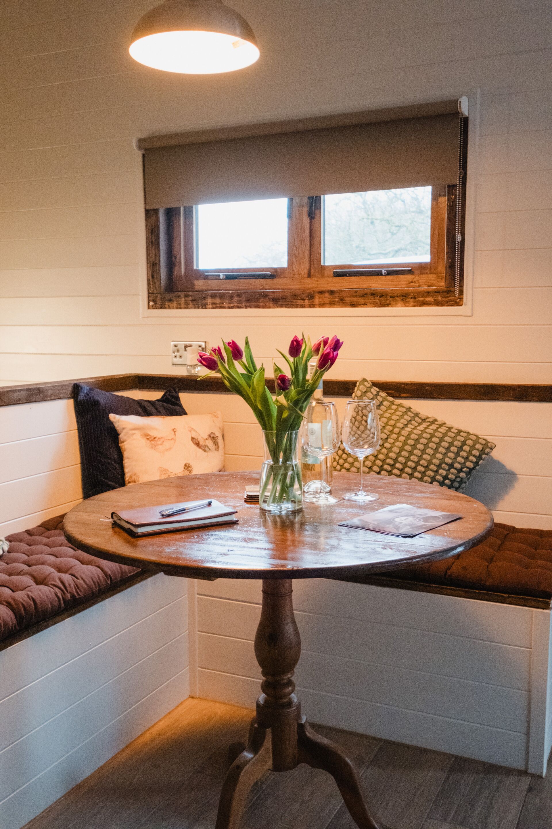 A corner seating area with a round table, a vase with flowers, a notebook, and a glass.