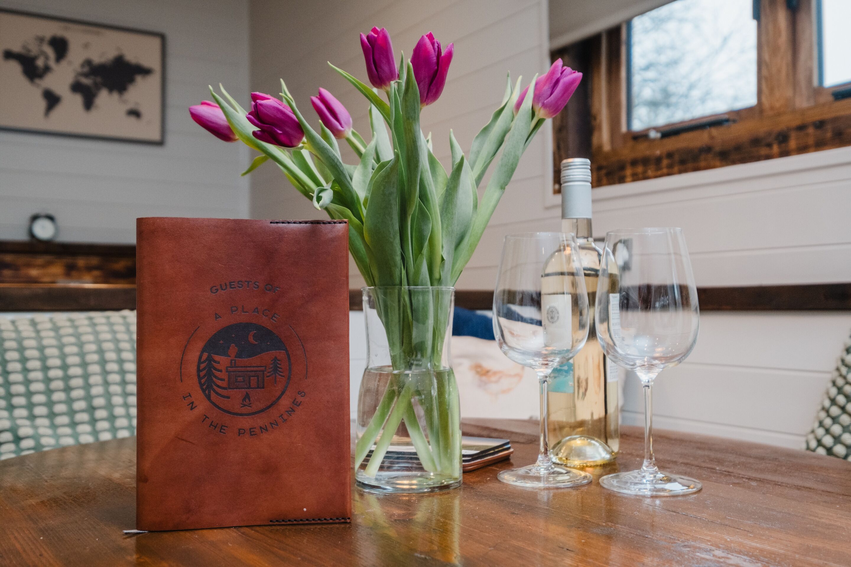 Un tavolo di legno con una borsa in pelle marrone, un vaso con tulipani viola e due bicchieri da vino vuoti.