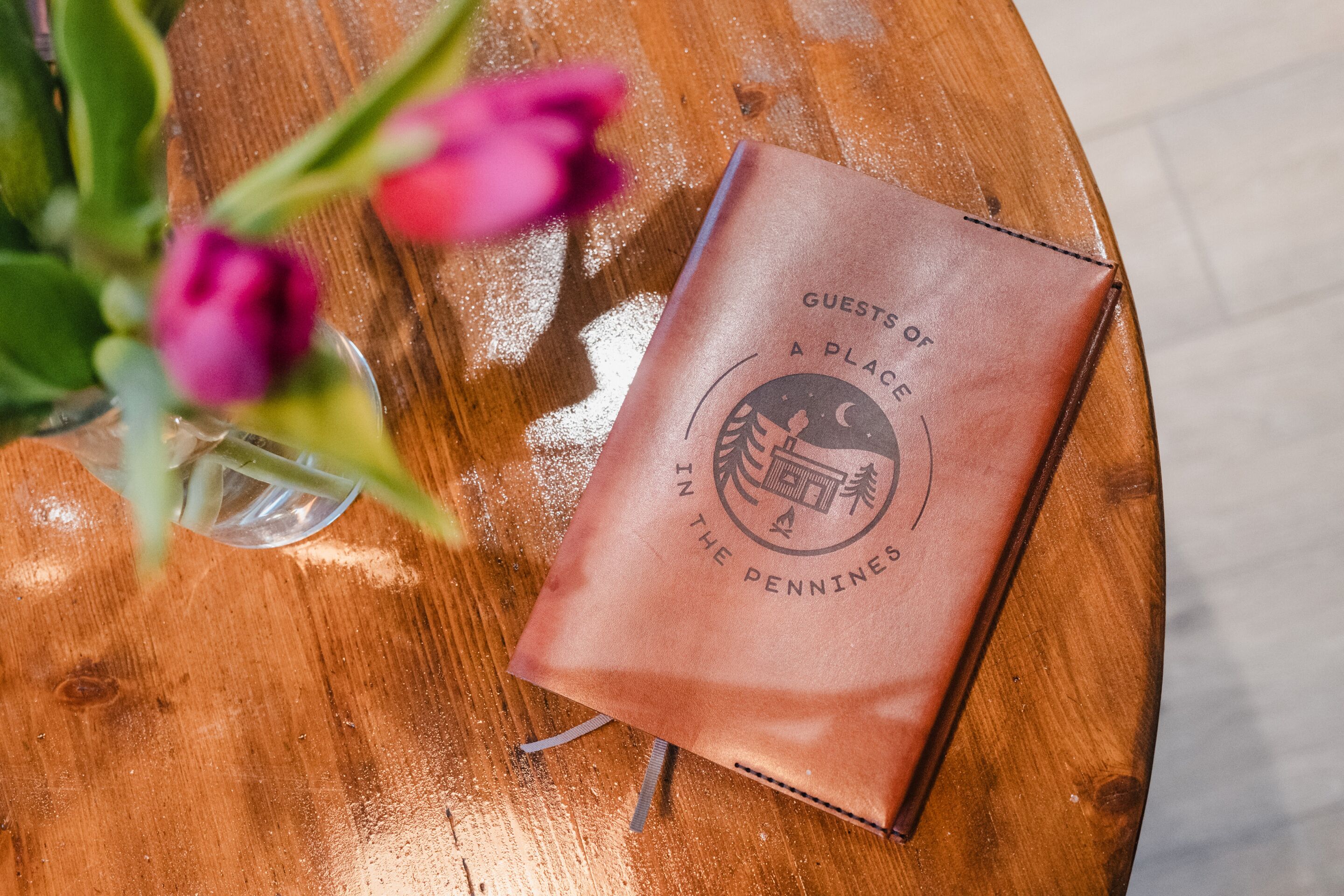 A brown leather-bound book titled "GUESTS OF A PLACE IN THE PENNINS" on a wooden table.