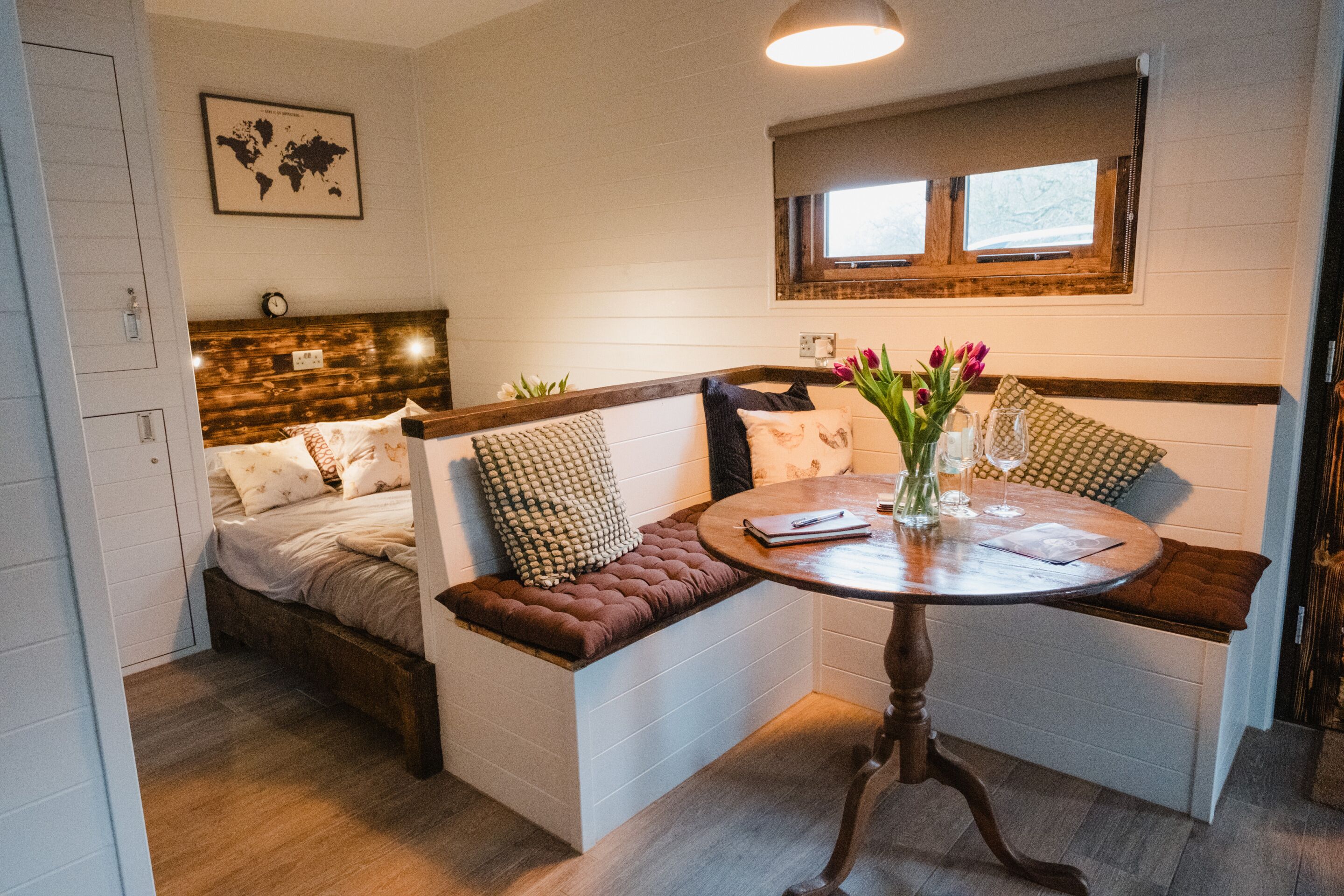 A cozy room with a bed, a bench with cushions, a round table with a vase of flowers, and a world map on the wall.