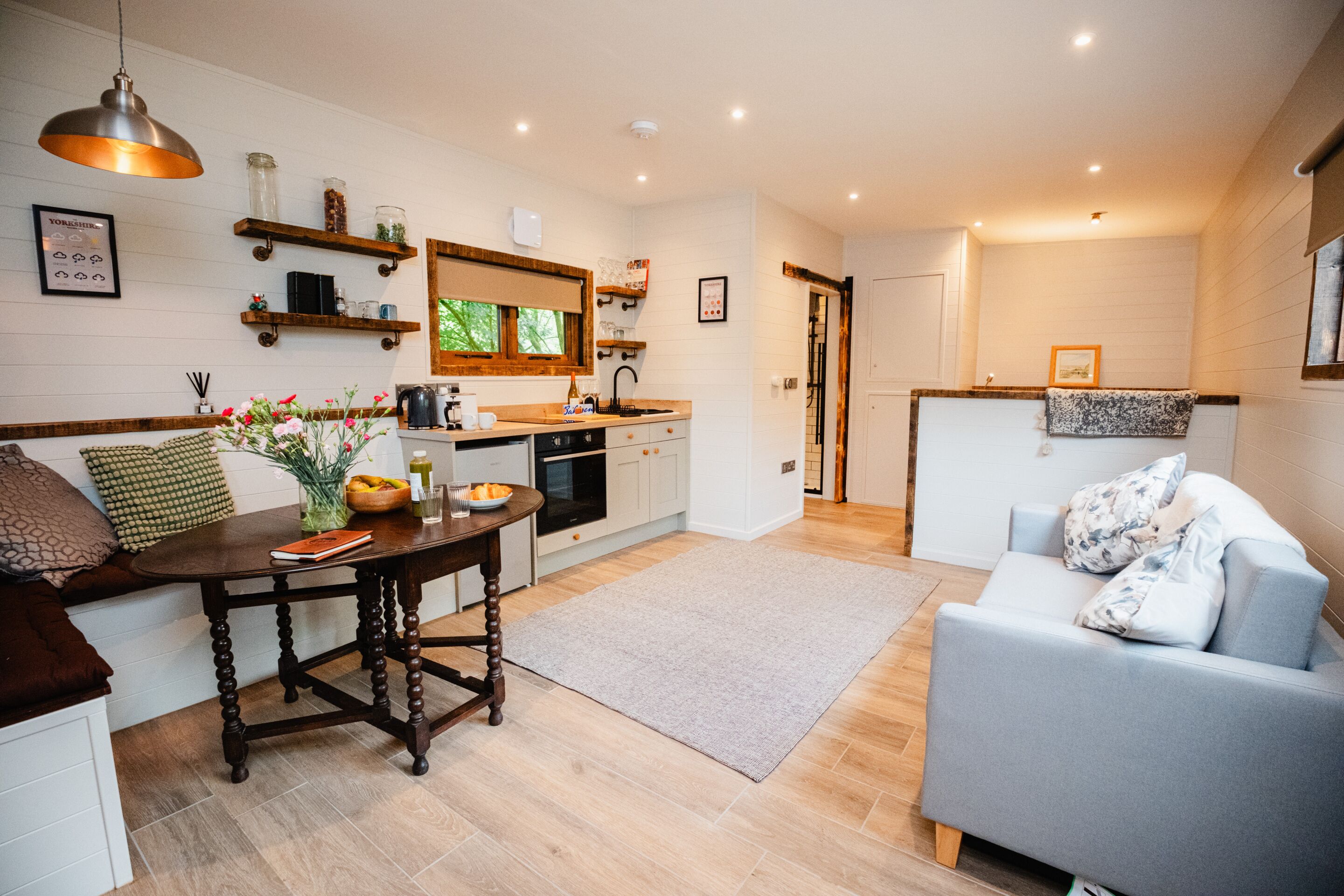 A modern kitchen with wooden shelves, a dining table, and a grey sofa in the living area.