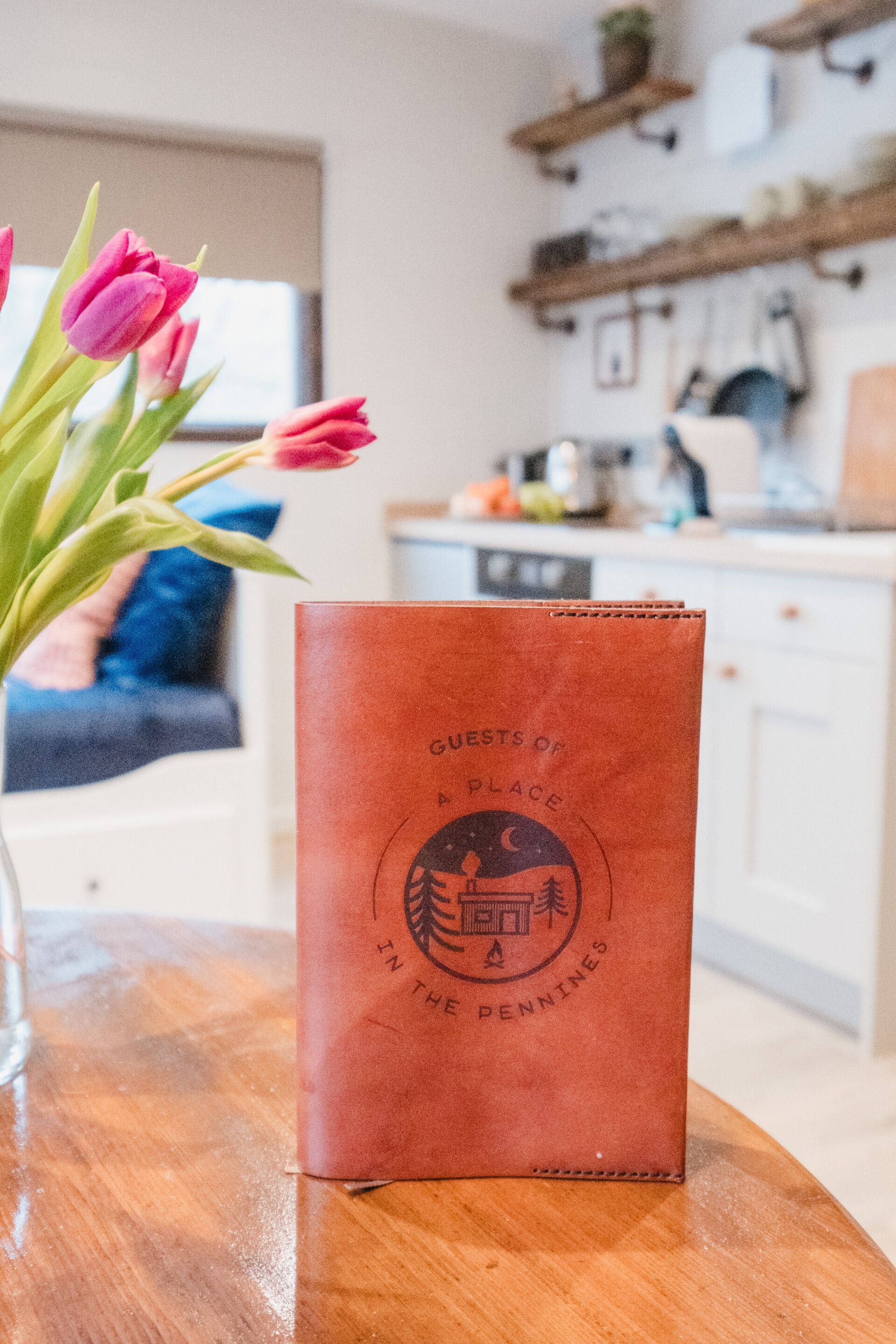 Una mesa de madera con un bolso de cuero marrón con la etiqueta "Guests of A Place in the Pennines" y un jarrón con tulipanes rosados.