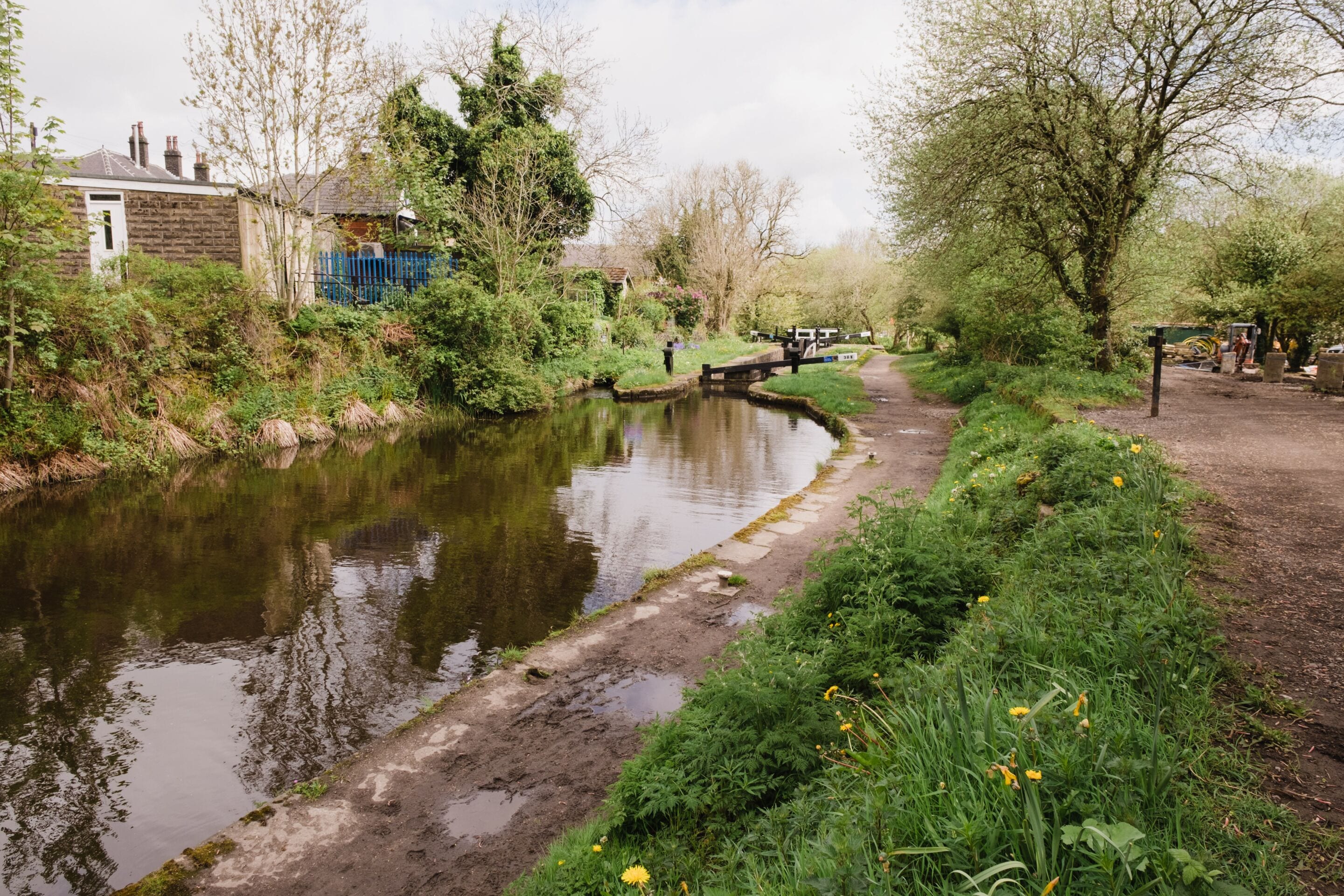 A narrow waterway with a wooden bridge, surrounded by greenery and a dirt path.