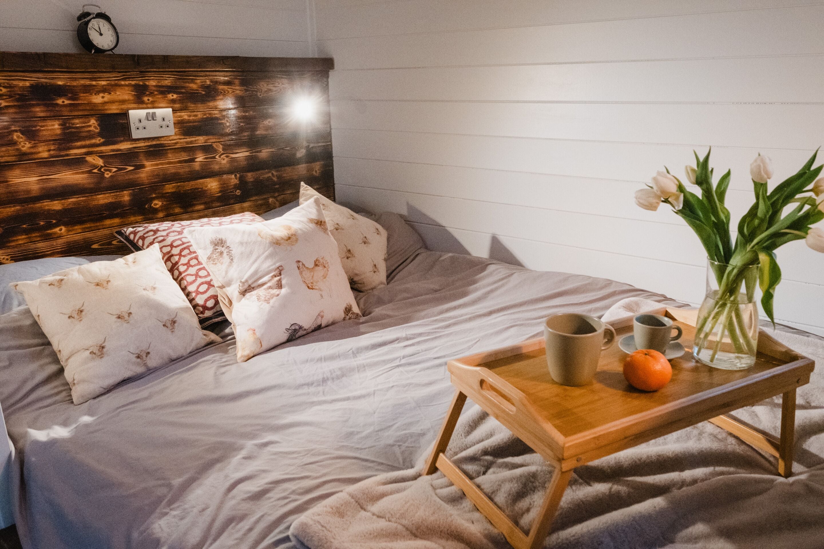 A neatly made bed with patterned pillows, a bedside table with a tray holding a mug, a small orange, and a vase with flowers.