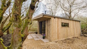 Une cabane en bois avec une porte et une fenêtre, entourée d’arbres et de gravier.