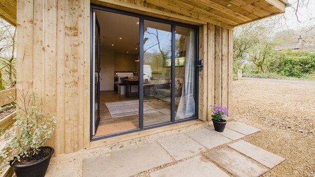 Un bâtiment en bois doté d’une porte coulissante en verre qui donne sur un espace intérieur comprenant un coin repas et un canapé.
