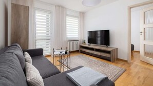 Una sala de estar moderna con un sofá gris, un televisor sobre un mueble de madera, una mesita con una planta y un ventanal grande con persianas.