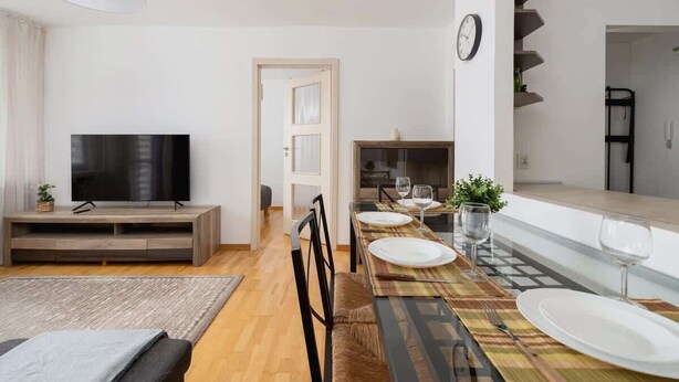 Una sala de estar moderna con un televisor de pantalla plana, un mueble de entretenimiento de madera y una zona de comedor con una mesa dispuesta para dos.