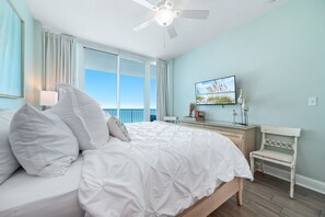 Une chambre avec un lit, un ventilateur de plafond, une fenêtre avec des rideaux, un bureau avec une chaise et un téléviseur fixé au mur.