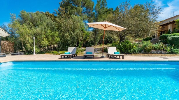 En klarblå pool med solstolar och ett parasoll, omgiven av frodig grönska och en byggnad i bakgrunden.