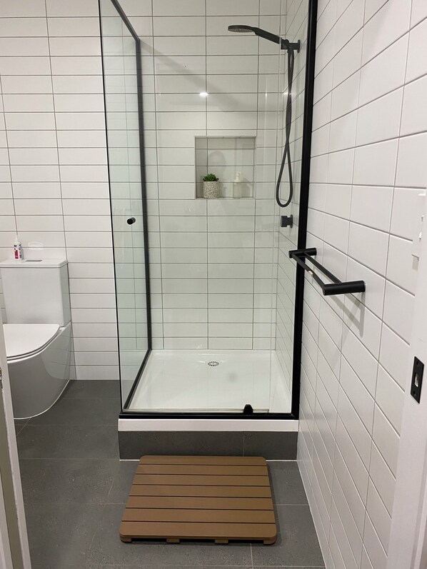 A glass-enclosed shower stall with a black frame, a small shelf with a plant and a bottle, and a wooden bench at the bottom. - Self-contained Studio in Pimpama - Gold Coast. Close to all major theme parks. (Pimpama)