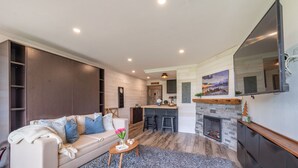 Een moderne woonkamer met een bank, salontafel en open haard. Achtergrond toont een keuken met een bar en krukken.